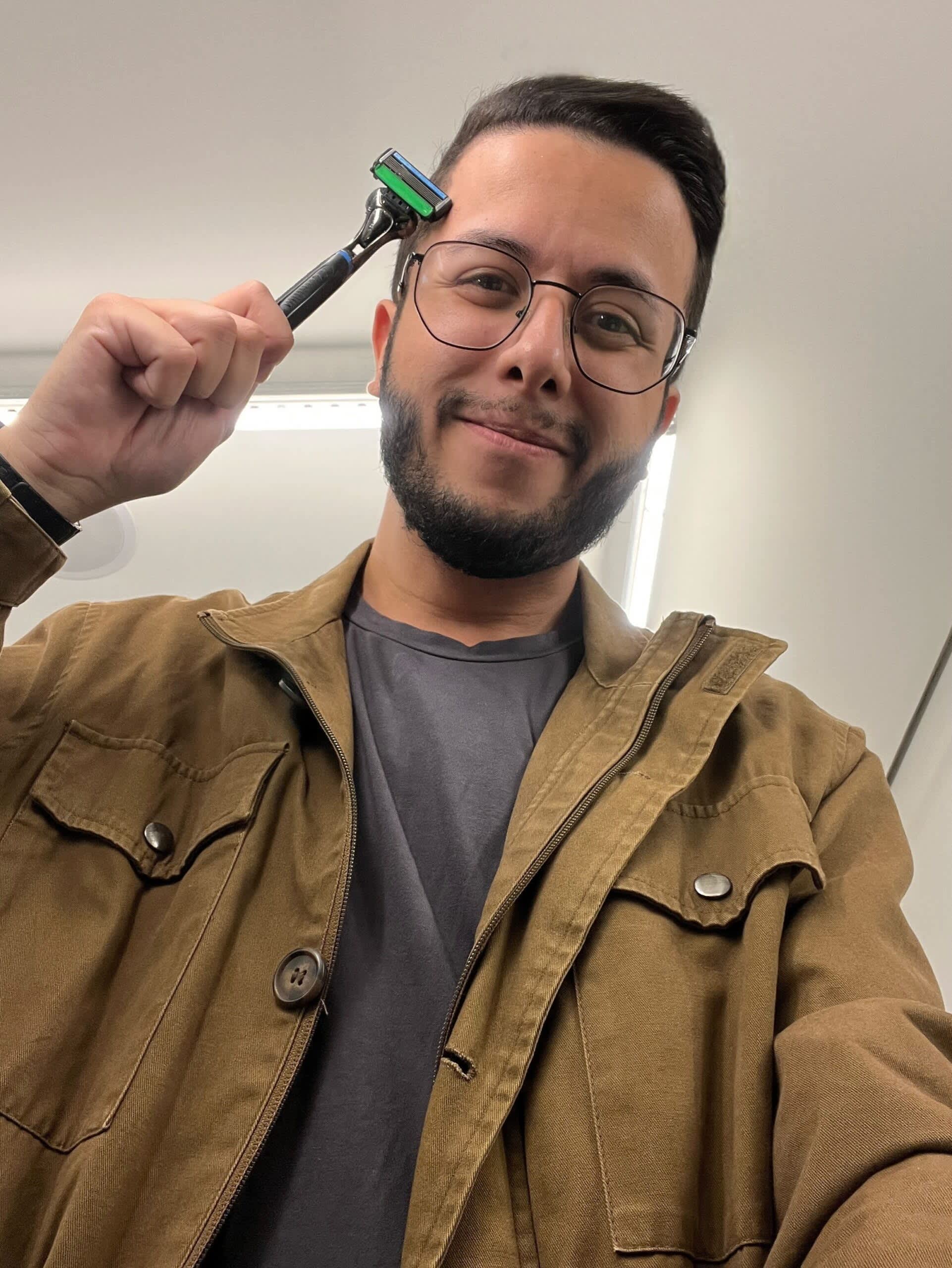 Um homem sorridente de óculos e barba, vestindo uma jaqueta marrom e camiseta cinza, segura um barbeador acima de sua cabeça.