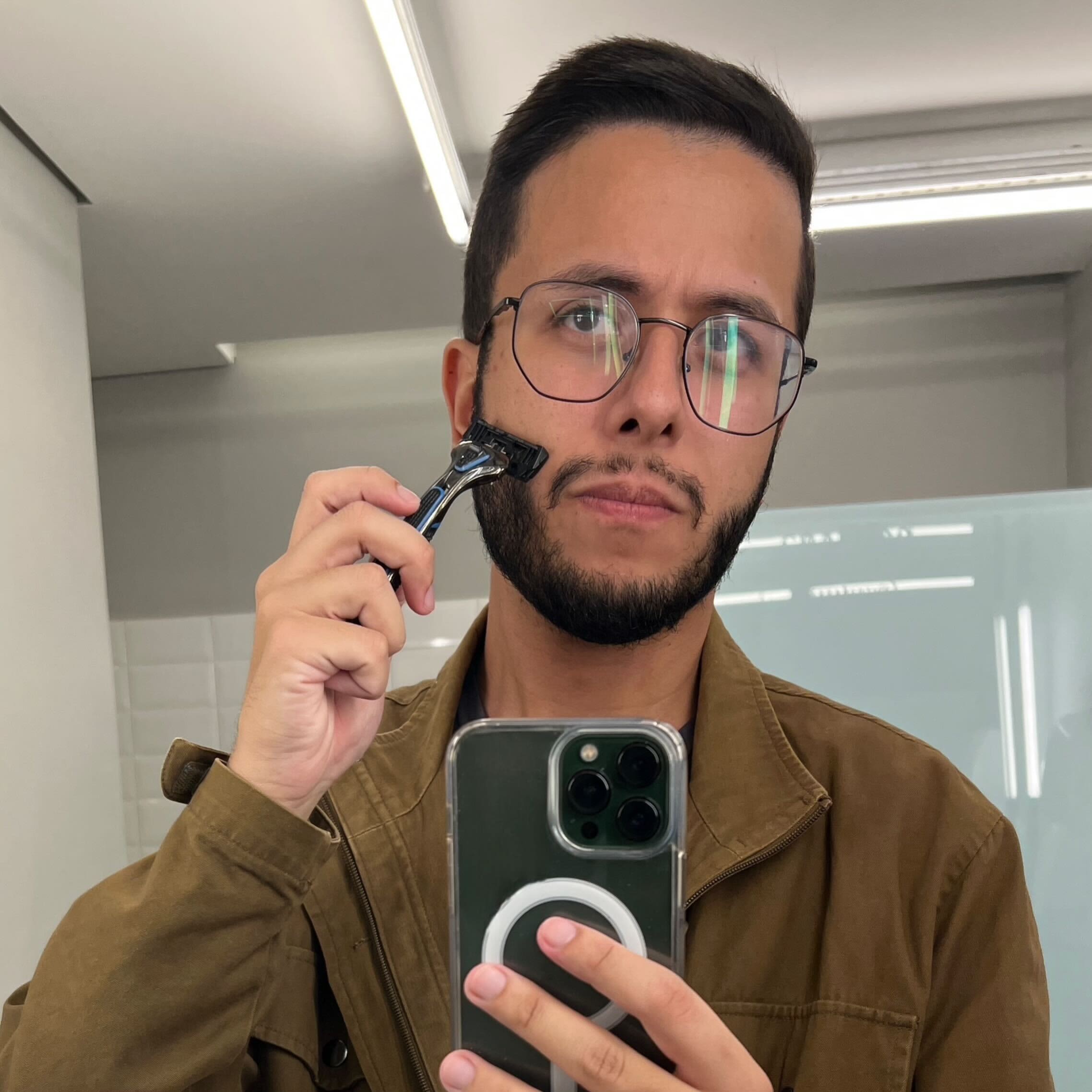 Um homem com barba e óculos está se barbeando com um barbeador manual, olhando para um espelho, enquanto se fotografa com um smartphone. Ele veste uma jaqueta marrom.