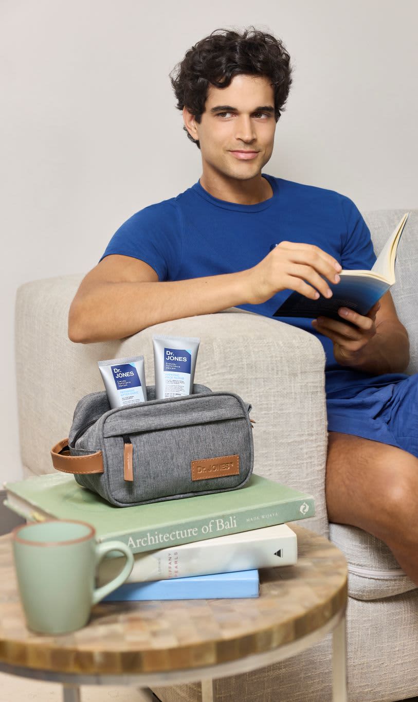 Homem sorrindo, sentado em poltrona, segura um livro. Ao lado, em uma pilha de livros, necessaire Dr. Jones com produtos. Mesinha com caneca. Ambiente claro e casual.