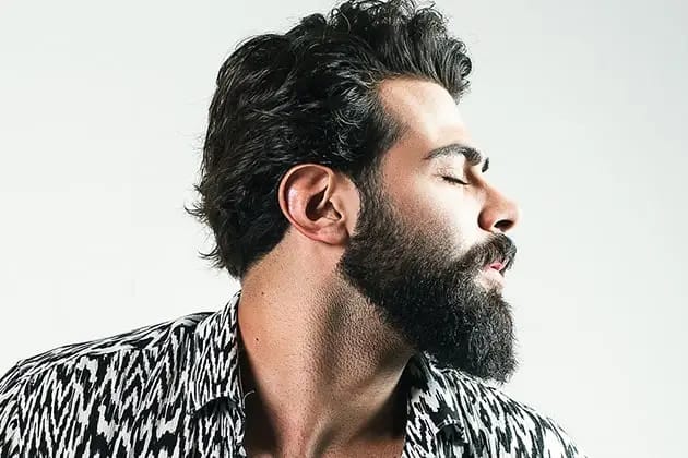 Em artigo sobre como tirar frizz da barba, imagem mostra homem de ascendência árabe e pele clara, barba quadrada e bem alinhada, de olhos fechados e cabelos escuros, virado para o lado direito. Ele usa camisa estampada preta e branca.