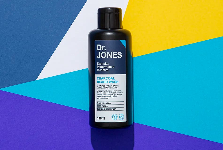 Foto do produto CHARCOAL BEARD WASH, da Dr. JONES. A embalagem do produto se encontra frente a um fundo geométrico nas cores azul-escuro, branco, amarelo, azul-claro e roxo. 