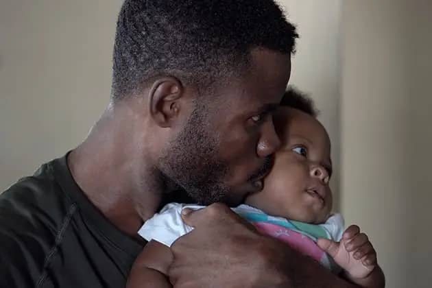 Foto de homem negro, cabelo raspado levemente e barba, camisa preta, segurando, abraçando e beijando seu bebê, que veste macaquinho branco, azul e rosa.