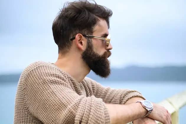 Em frente ao mar e apoiado em cerca branca, homem de blusa bege, relógio e óculos olha para o lado direito. Ele tem barba comprida e desenhada.