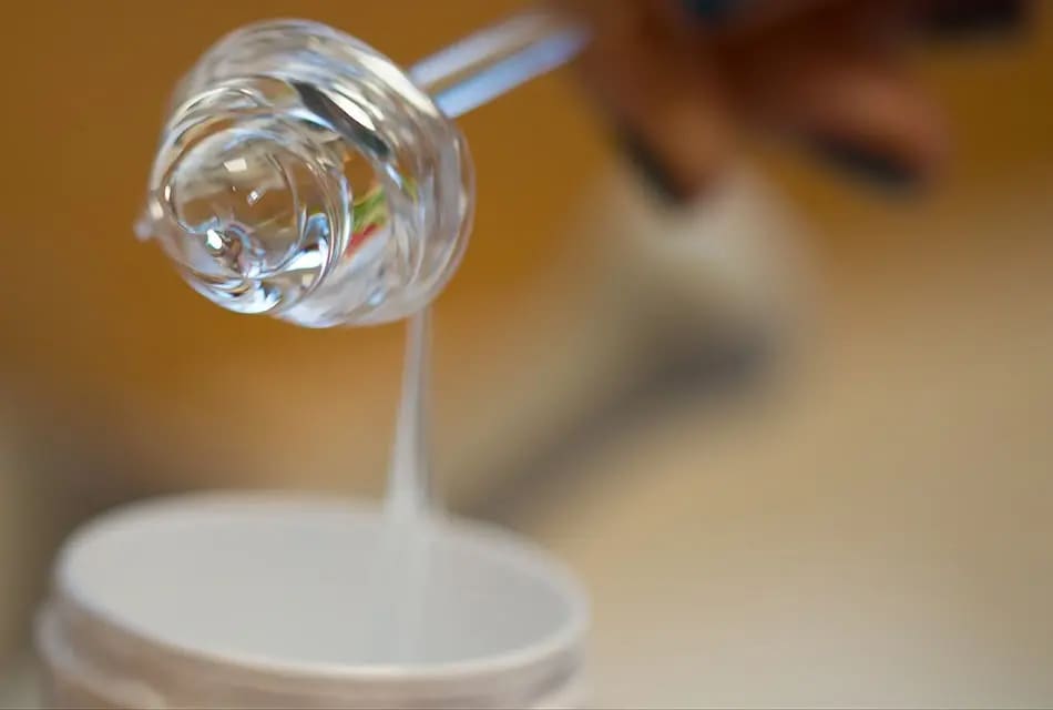 Imagem mostra pequena espátula em destaque, exibindo aspecto e viscosidade do Pantenol (Pró-Vitamina B5), que é um líquido quase incolor que escorre em direção a um tubo branco.