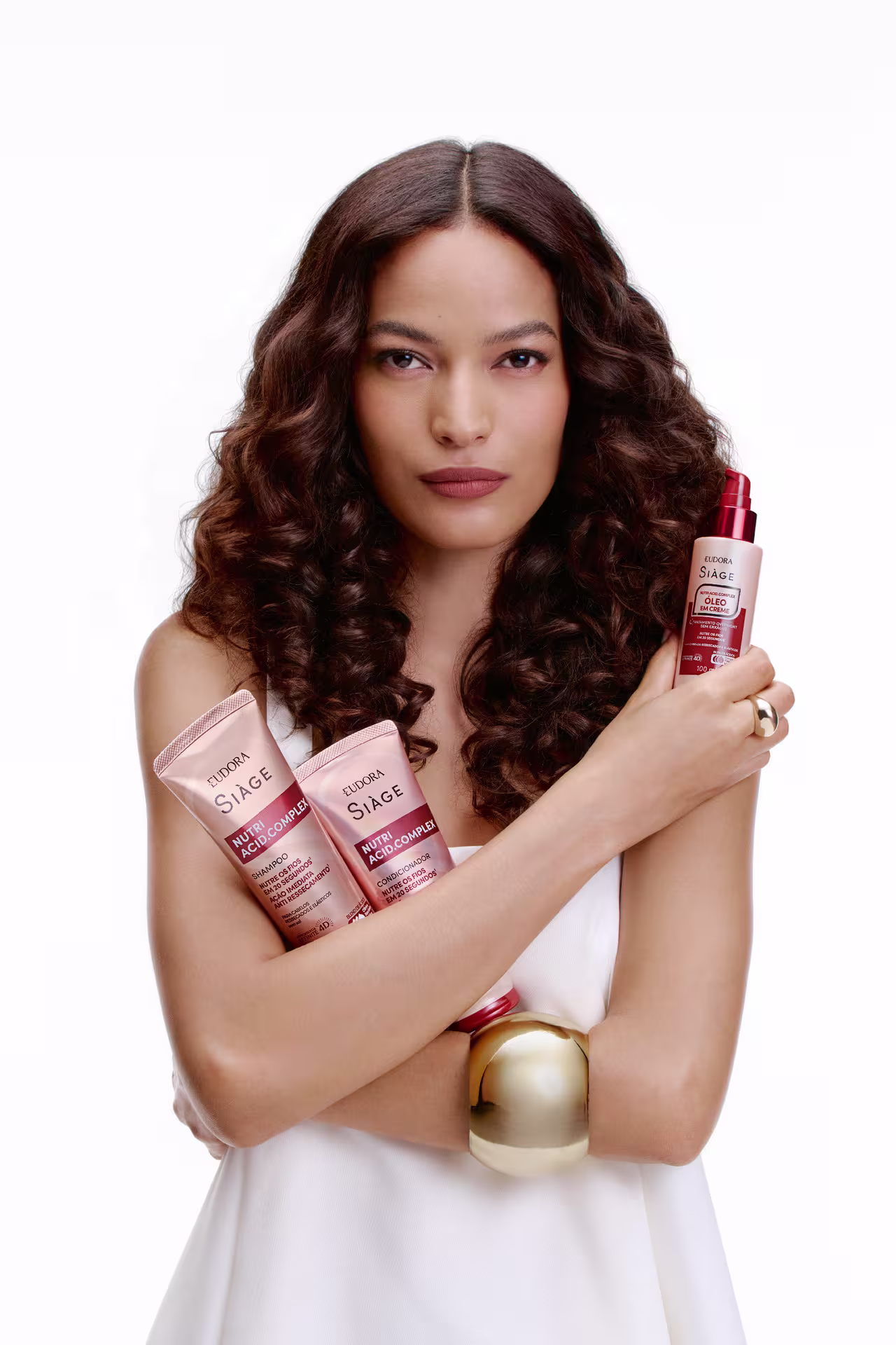 Mulher de cabelos castanhos ondulados e volumosos segura produtos da linha Eudora Siàge Nutri Acid. Complex. Usa roupa branca, bracelete dourado grande e exibe maquiagem neutra elegante.