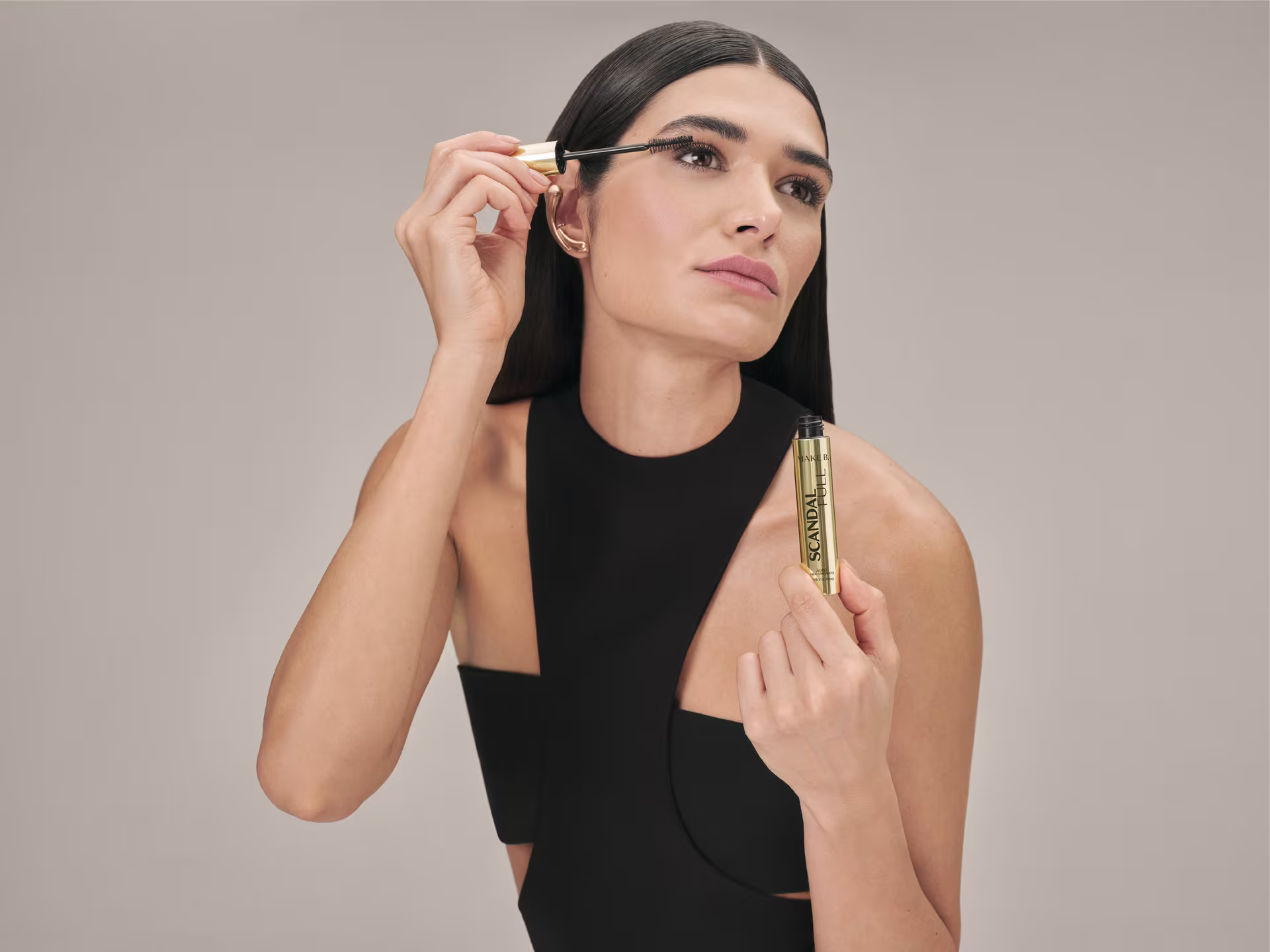 Mulher de cabelos escuros e lisos aplica máscara de cílios Make B. Scandal Lash com o pincel aplicador, enquanto segura o frasco dourado na outra mão. Ela usa roupa preta e o fundo é bege neutro.