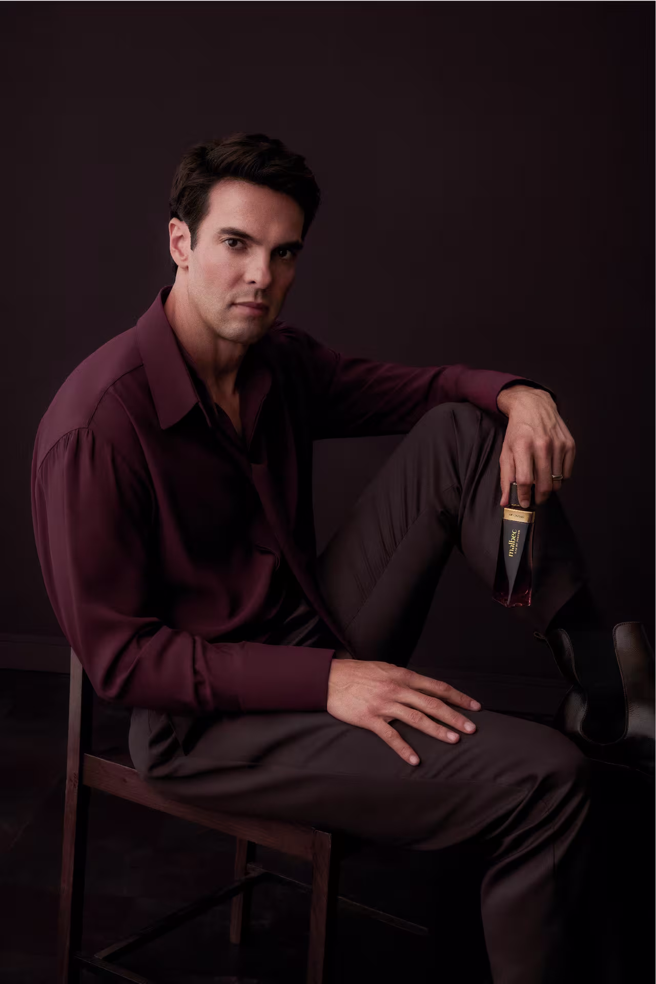 Kaká sentado em uma cadeira de madeira, vestindo camisa vinho, segurando o frasco da fragrância Malbec em um fundo escuro.