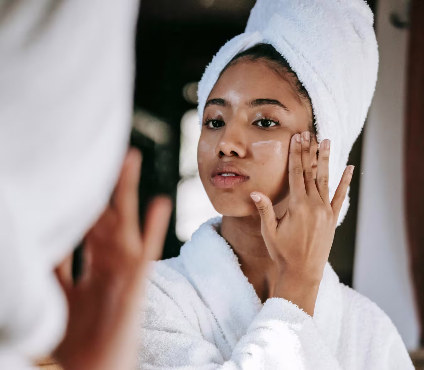 Mulher fazendo skincare para pele oleosa: ela está de toaolha branca na cabeça e de roupão. também branco. Ela está olhando o reflexo no espelho e aplicando um creme no rosto.