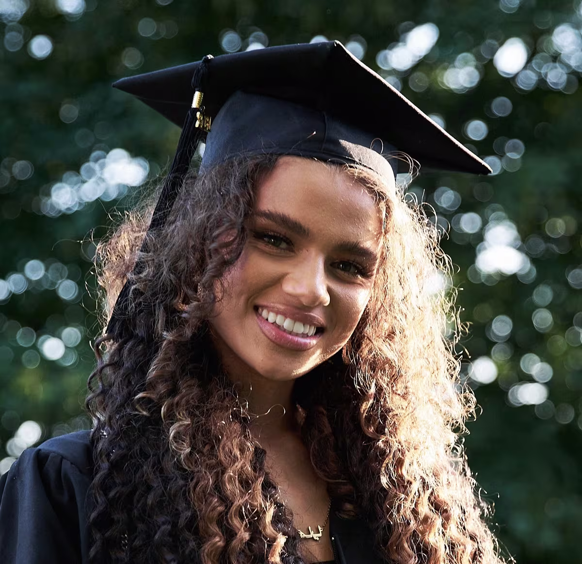 Mulher negra clara, jovem com cabelos longos cacheados. Ela está sorrindo para a foto e veste beca e chapéu de formatura, ambos de cor preto.