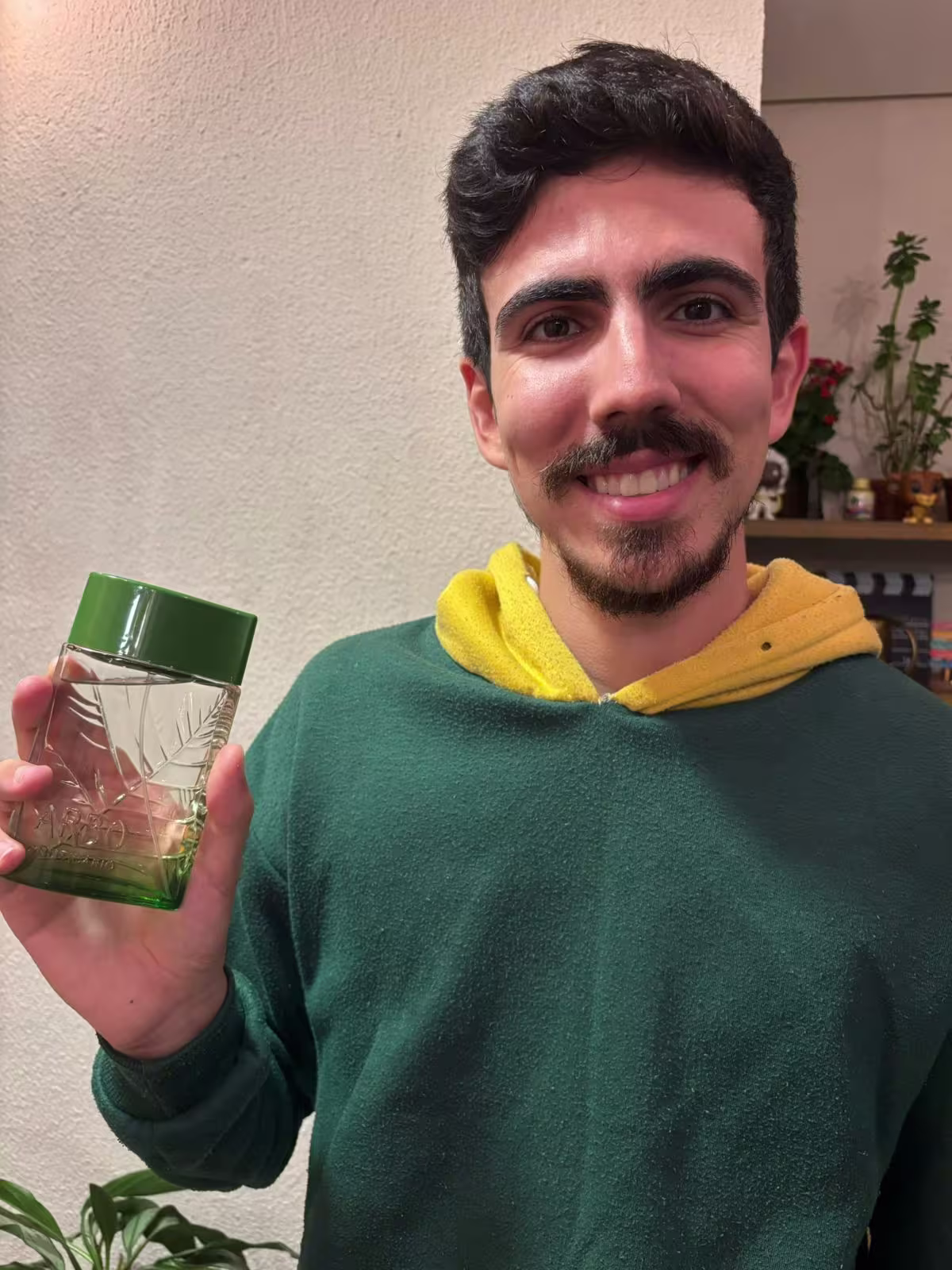 Um homem jovem e sorridente segura um frasco de Arbo Puro. Ele está vestindo uma camisa verde escura