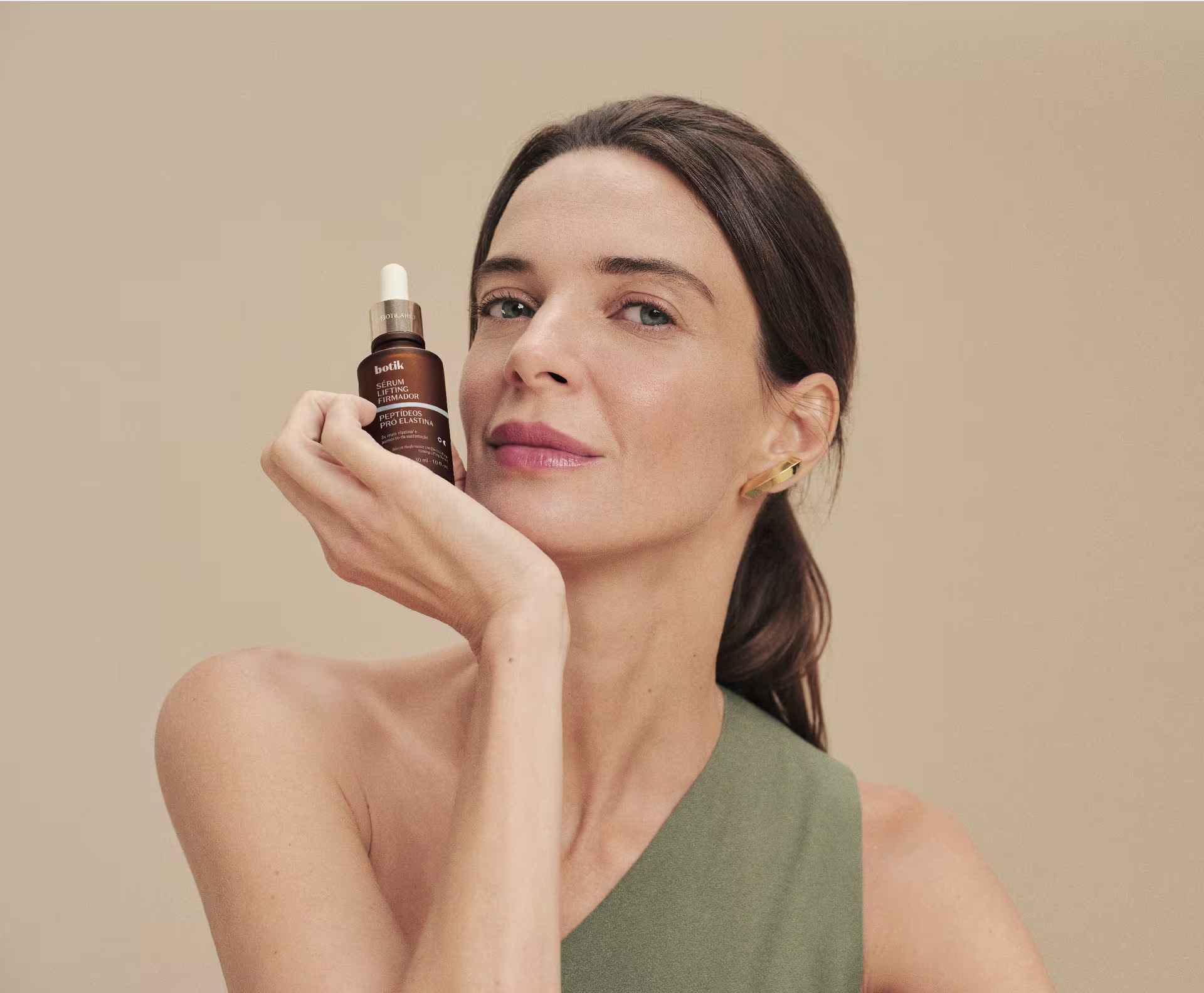 Mulher de cabelos presos e blusa verde sorri levemente, apoiando o rosto na mão enquanto segura um frasco conta-gotas do sérum Botik sobre um fundo neutro.