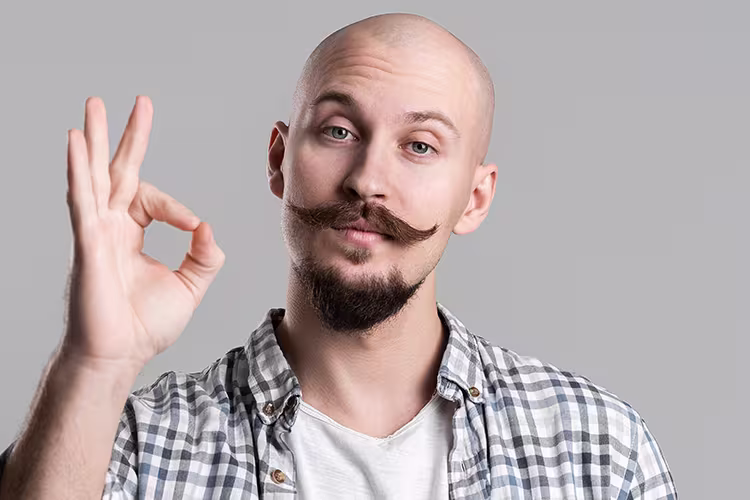 Barba para carecas: Homem careca branco jovem de bigode estilo 'guidão de bicicleta' e cavanhaque sinaliza para a câmera. Ele usa camiseta branca e blusa quadriculada.