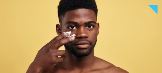Jovem negro de cabelos curtos e barba por fazer passa creme em seu rosto, em frente a fundo amarelo.