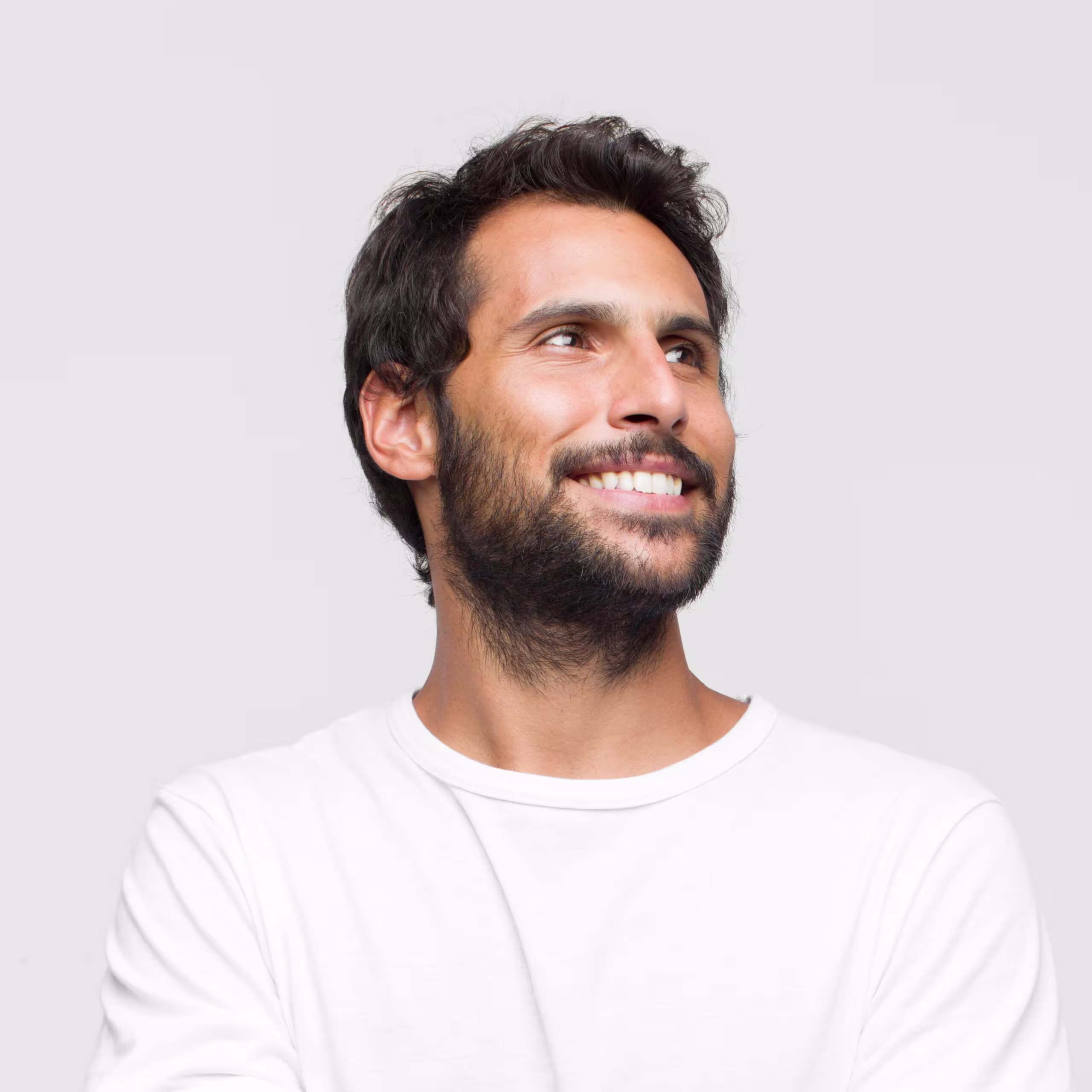 Homem jovem de barba e cabelos ondulados sorrindo e olhando para o lado, usando camiseta branca, com fundo cinza claro. Dicas de visagismo masculino