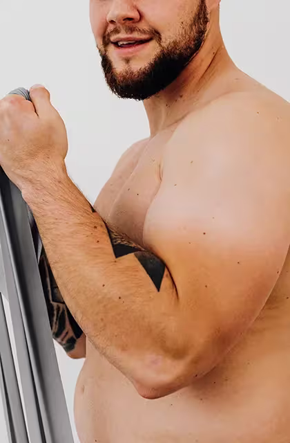 Foto de um homem forte e branco, de barba, recortada abaixo dos olhos. Ele está tirando a camisa e exibe o peito e uma barriga mais saliente, tem tatuagem no antebraço, usa barba e está de perfil.