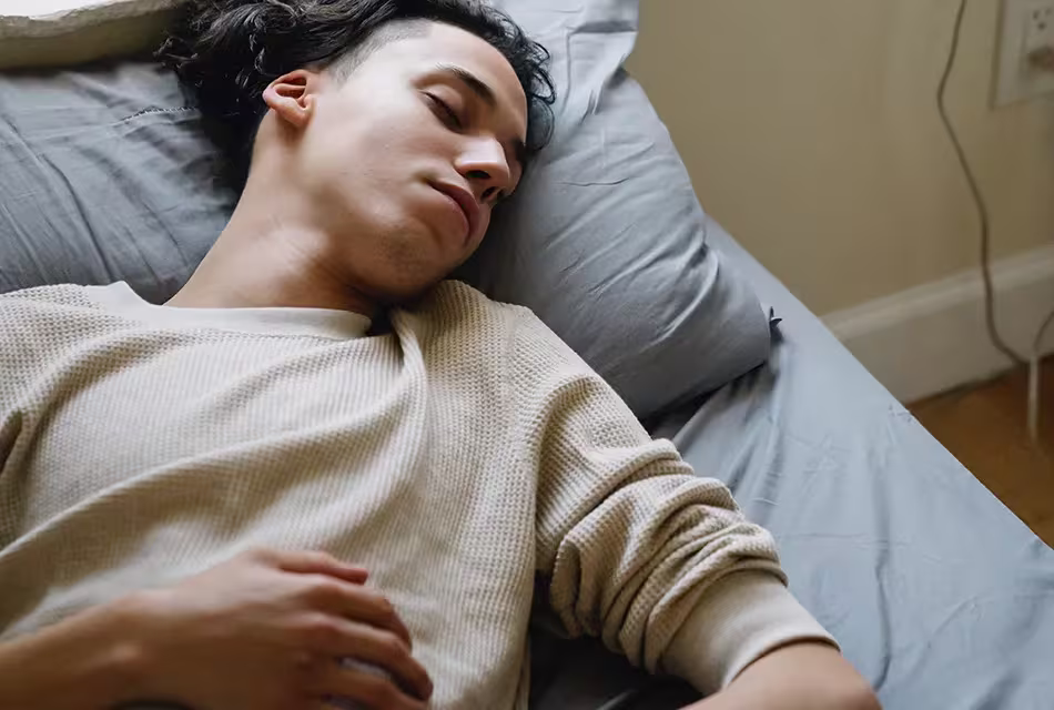 Jovem de pele clara e cabelos escuros e médios, raspados na lateral, dorme em cama de colcha e travesseiros azuis, usando blusa bege.