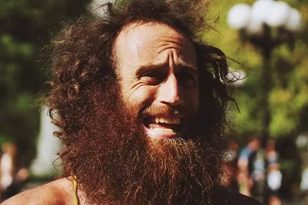 Em artigo sobre frizz na barba ('barba crespa'), imagem mostra homem fazendo careta, pele clara, barba e cabelos bem desgrenhados e grandes, com bastante frizz, em meio a uma praça.