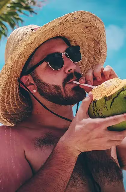 De chapéu de palha, rapaz sem camisa, pele clara e óculos escuros, bebe água de coco utilizando um canudo, embaixo de coqueiro.