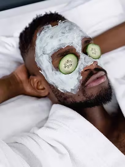 Homem negro está de robe branco em cima de uma cama, deitado, mãos atrás da cabeça, com creme no rosto e olhos cobertos com rodelas de pepino. Ele tem cabelos curtos, raspados na lateral, e usa barba.