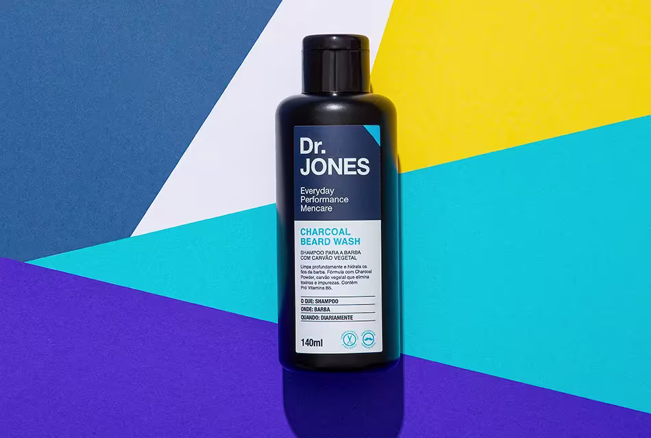 Imagem da embalagem do Charcoal Beard Wash, da Dr. JONES, com Carvão Vegetal ativado, em fundo geométrico colorido nas cores azul-claro, azul-escuro, roxo, branco e amarelo.