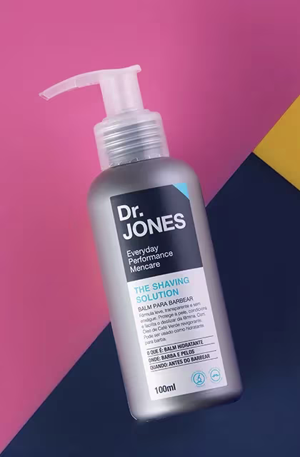 Em fundo com recortes em rosa, azul e amarelo, foto em destaque da embalagem do balm para barbear The Shaving Solution, da Dr. JONES.