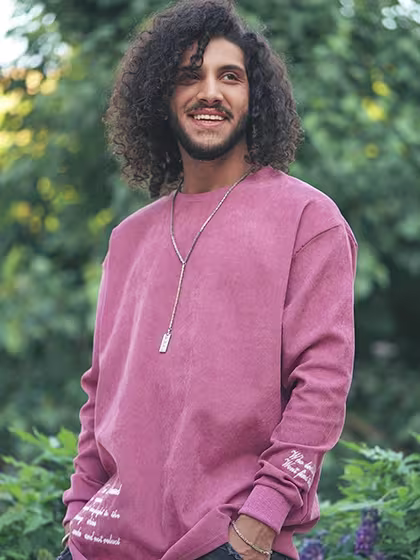 Foto de homem pardo de cabelos médios encaracolados e barba aparada. Ele sorri e usa calça jeans e blusa rosada, para aludir ao tratamento bem-sucedido com o climbazol em shampoos anticaspa.