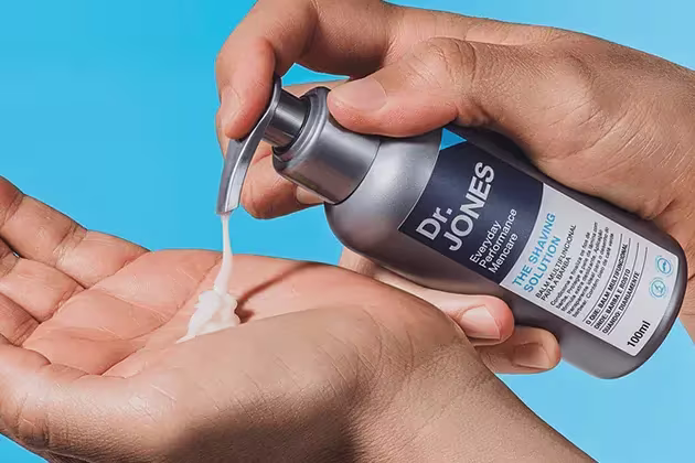 Foto que mostra um usuário pressionando embalagem do balm para barba The Shaving Solution, da Dr. JONES, e o conteúdo despejado na outra mão.