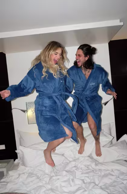 Thaigo Consani e Gabi Domingues, de roupões azuis, pulam em cama com lençóis brancos na suíte Dr. JONES no Hotel Unique.