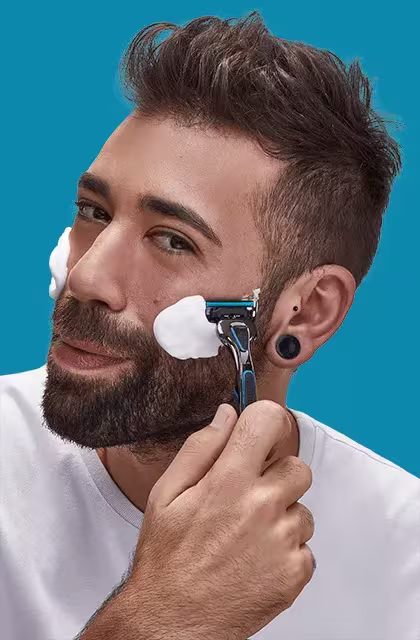 Em fundo azul, homem de pele clara, barba curta, olhos castanhos e cabelo com topete e raspado nas laterais, utiliza o barbeador THE RAZOR4, da Dr. JONES, para fazer o detalhe da bochecha, na qual há aplicação da espuma de barbear PRECISION FOAM.