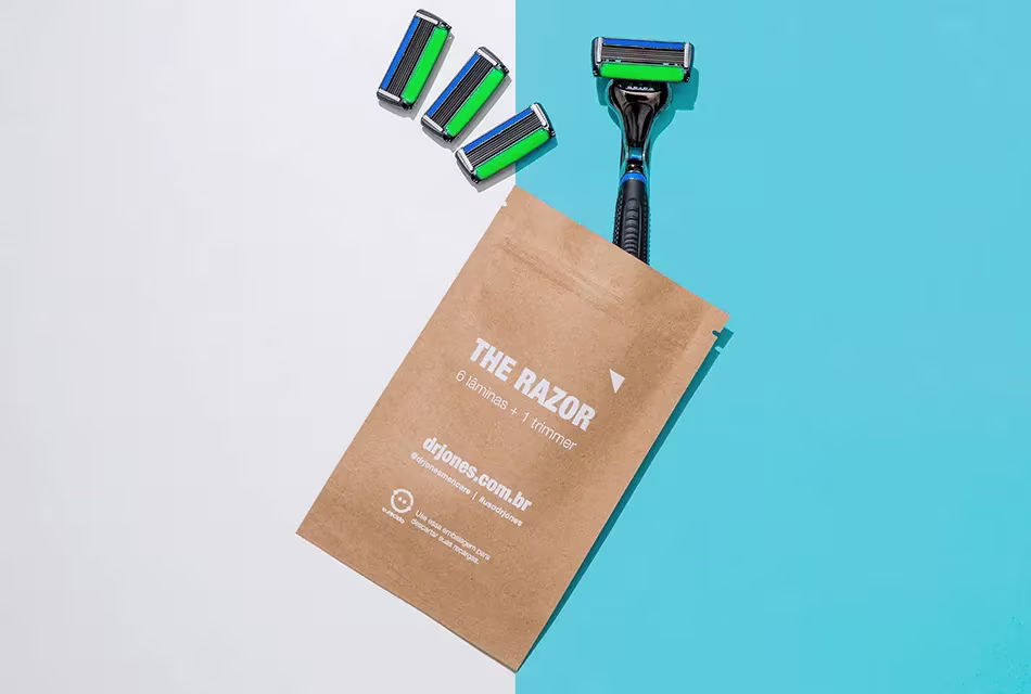 Foto de envelope em papel pardo com o THE RAZOR6 e quatro recargas do barbeador da Dr. JONES. O envelope destina-se ao descarte das lâminas. O fundo é cinza e azul.