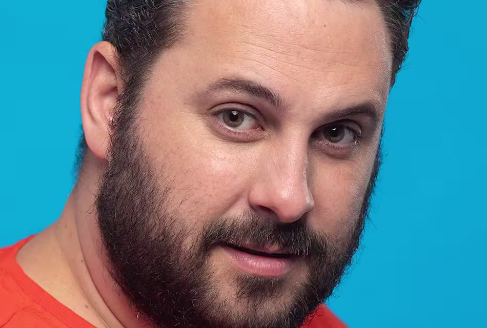 Homem gordo, de camiseta laranja, de pele clara, olhos verdes e barba curta e densa olha para a câmera em frente a fundo azul.