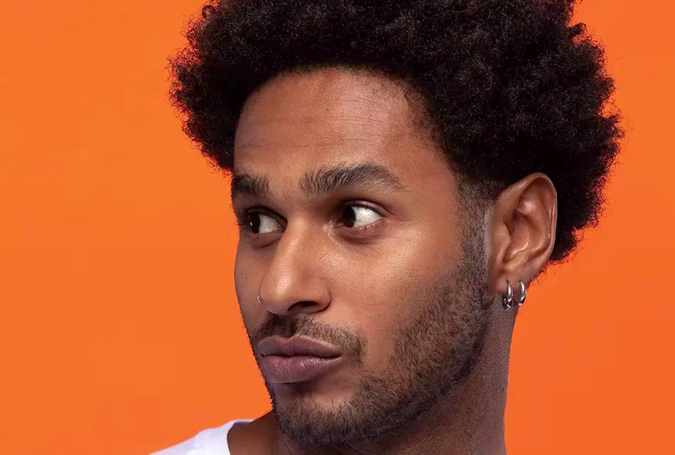 Homem jovem e negro, de cabelos em estilo black power, olhada para o lado em frente a fundo laranja. Ele tem a barba por fazer e usa dois brincos na orelha.