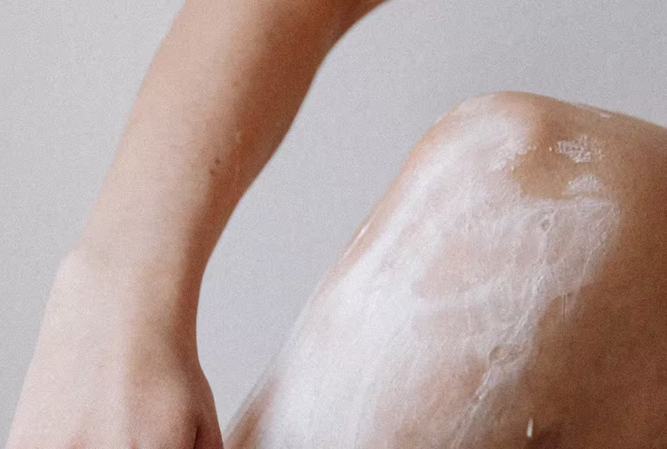 Foto de perna feminina em que se vê, em destaque, o joelho e a região próxima com espuma de barbear ou creme, preparando a região para a depilação.