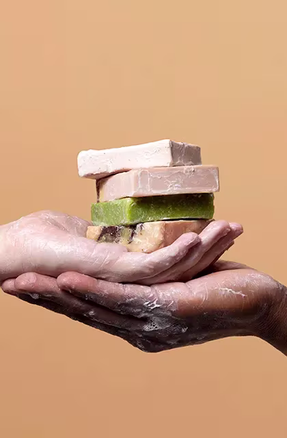 Close de mãos, uma branca e uma negra, uma embaixo da outra, segurando quatro sabonetes nas cores bege, rosa, verde e laranja, em fundo laranja.