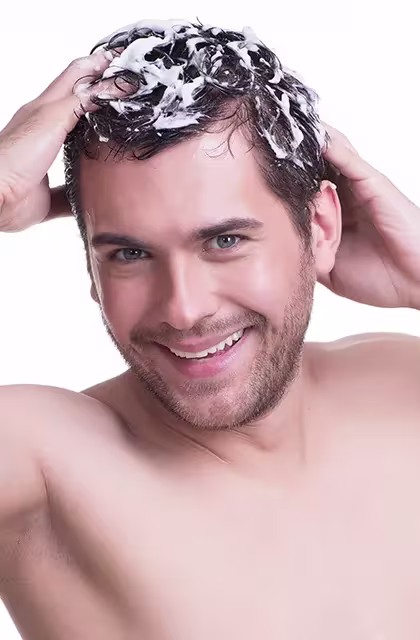 Homem de pele clara, barba por fazer e olhos claros, sem camisa, passando shampoo nos cabelos escuros.
