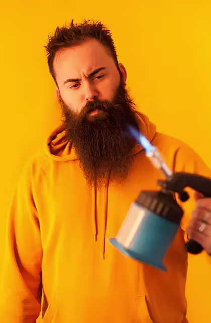 Foto de homem com agasalho amarelo, barba lenhador, cabelos curtos e fundo amarelo segurando um maçarico.