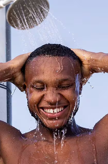 Jovem negro sorri enquanto toma banho em chuveiro externo, localizado na praia. Ele tem cabelos curtos e barba por fazer nas laterais, sem bigode.