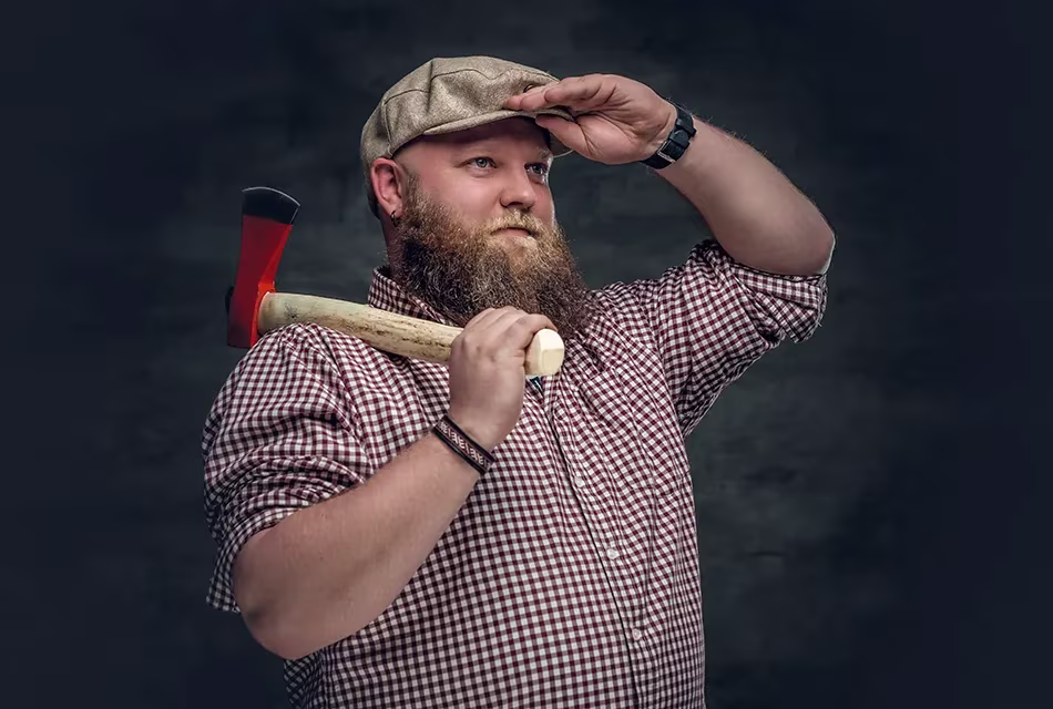 Retrato de homem de pele clara, olhos azuis, acima do peso, com barba lenhador, camisa quadriculada e um boné sobre fundo cinza segurando um machado e com as mãos na testa em sinal de sentido.