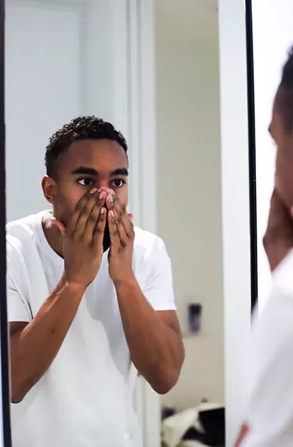 De camiseta branca, jovem negro de cabelos cacheados e curtos, raspados nas laterais, passa creme olhando para o espelho. Ele põe as mãos em torno do nariz, finalizando os cuidados com a pele.