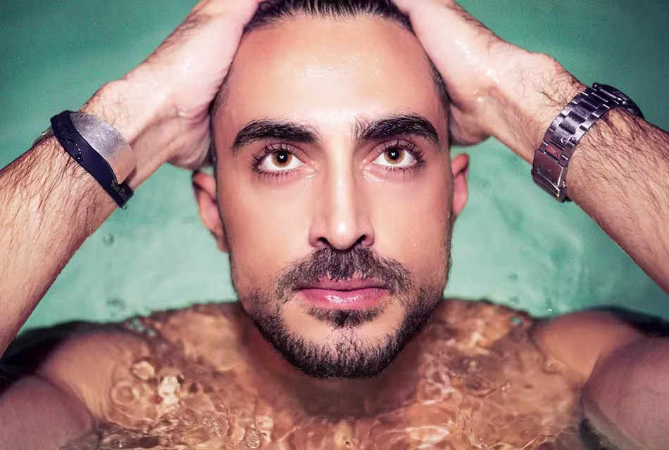 Homem de pele clara, cabelos escuros e olhos castanho-esverdeados, barba por fazer, sai da piscina, olhando para cima, com as mãos nos cabelos para retirar a água do rosto. Ele usa num dos pulsos um relógio e em outro uma pulseira.