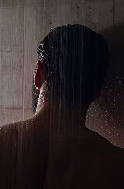 De costas, vê-se, à meia-luz, homem de barba média e cabelos escuros curtos e lisos sob água do chuveiro.