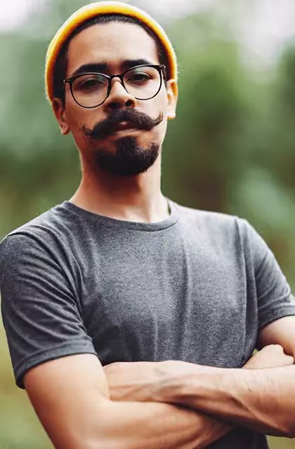 Rapaz pardo, de óculos arredondados e camiseta cinza, olha para a câmera, de braços cruzados. Ele é pardo, usa um gorro laranja e bigode handlebar com cavanhaque denso.