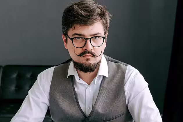 Homem de pele clara usa colete cinza com camisa social. Os cabelos são curtos, ele tem um cavanhaque denso e um bigode handlebar longo.