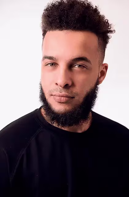 Rapaz negro de olhos cor de mel e camiseta preta olha para a câmera. Os cabelos e a barba, estilo Old Dutch, são castanho-escuros.