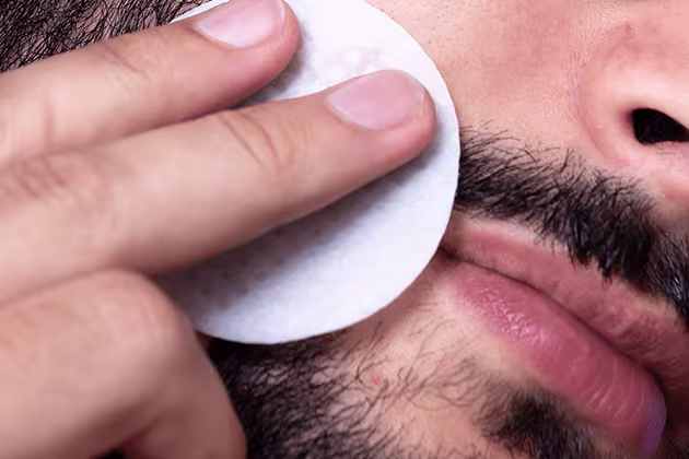 Foto em destaque mostra lateral de rosto masculino com barba, de homem de pele clara. Ele usa um algodão em forma de disco para fazer limpeza facial.
