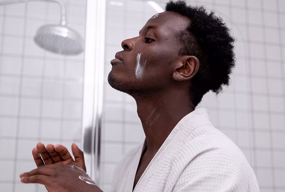 Foto de homem negro em banheiro, barba por fazer e cabelos curtos e raspados na lateral, passando creme no rosto e nas mãos. Ele usa um roupão branco e se olha em direção a um espelho. Atrás, vê-se o chuveiro.