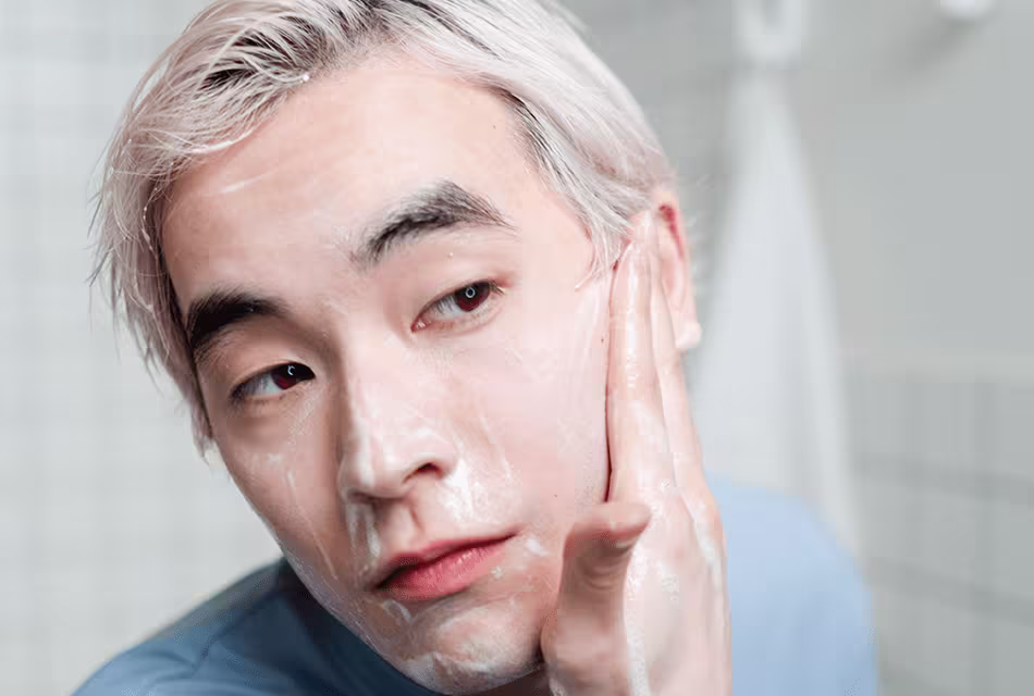 Foto de homem oriental e cabelos loiros passando creme no rosto e olhando-se em direção a um espelho. Ele é jovem, usa camiseta azul e não tem barba.