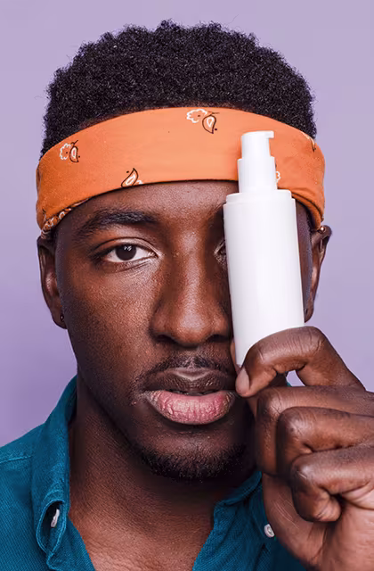 Homem negro e com bigode fininho e cavanhaque olha de frente para a câmera e segura produto de beleza com pump. Ele é jovem e usa faixa laranja na cabeça e camisa azul.
