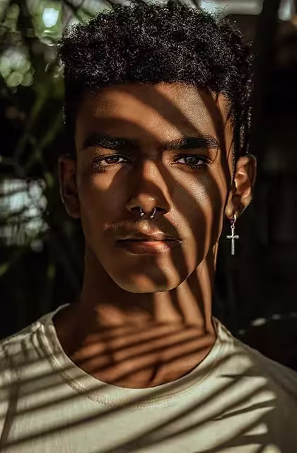 Foto de rapaz jovem e negro, cabelos curtos e camiseta branca, sem barba e brinco em formato de cruz. Ele se encontra embaixo de uma árvore, iluminado pelo sol, e olhando diretamente para a câmera.