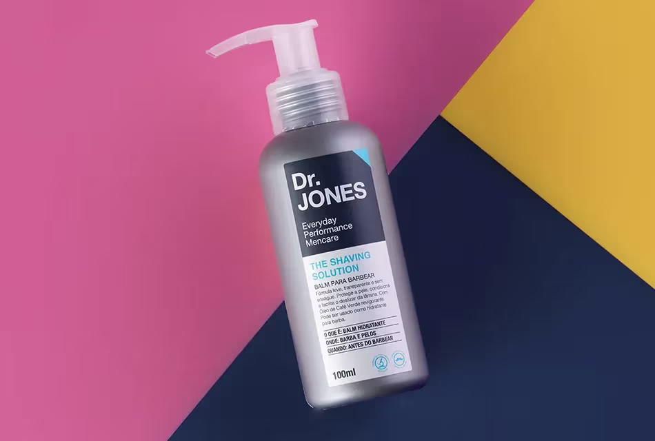 Foto da embalagem do balm The Shaving Solution, da Dr. JONES, em fundo rosa, azul-escuro e amarelo.